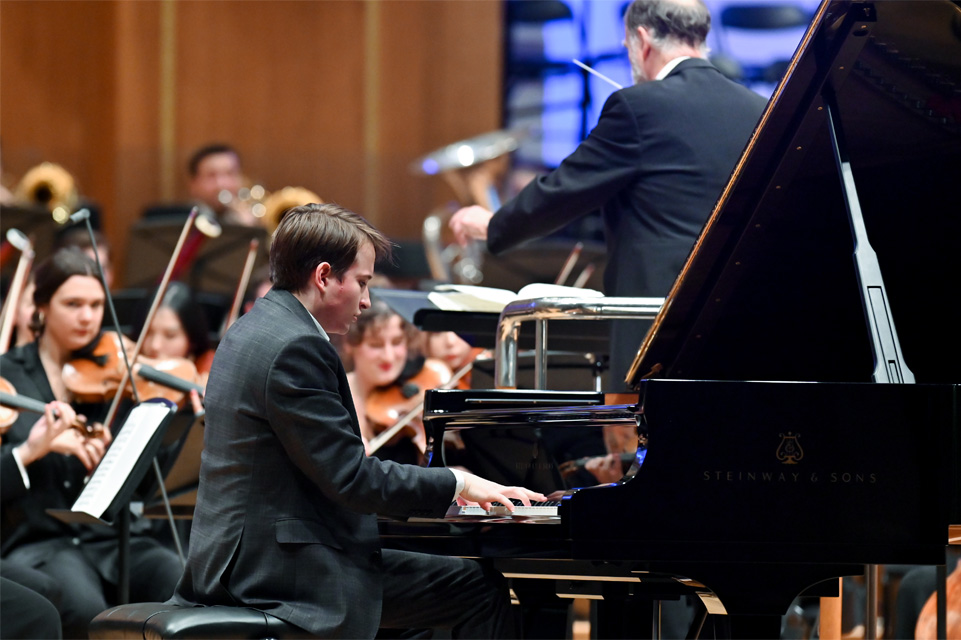 A male student playing the piano in an orchestra, with an orchestra being conducted in the background.